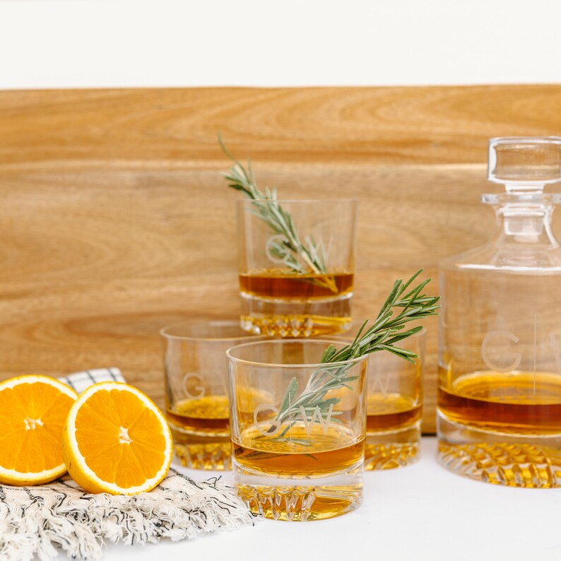 May include: Four clear glass tumblers with a decorative pattern on the bottom, filled with a golden liquid and garnished with sprigs of rosemary. A clear glass decanter with a stopper, also filled with the same golden liquid, sits on a wooden surface.