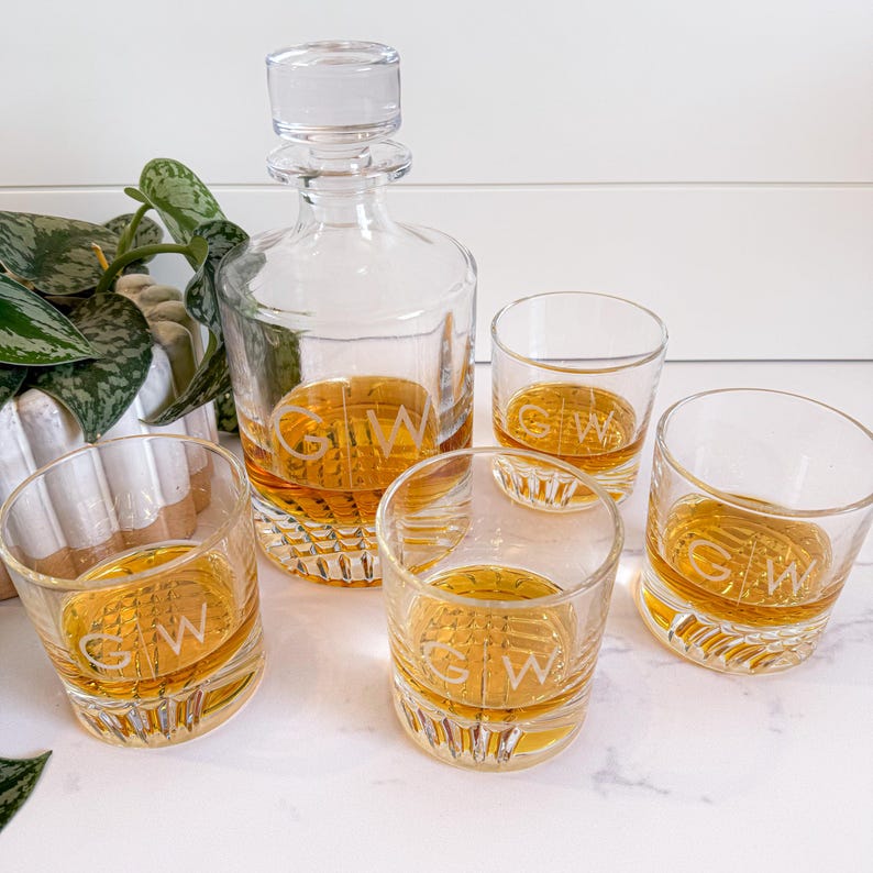 May include: A set of four clear glass whiskey glasses and a decanter, all with the initials "GW" etched on them. The glasses are filled with amber liquid.