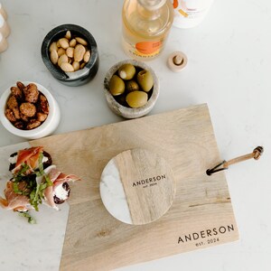 Puede incluir: Una tabla de servir de madera con un posavasos de m&aacute;rmol y madera con el texto "Anderson est. 2024". La tabla tiene una asa de cuero y est&aacute; rodeada de cuencos de aceitunas, nueces y un aperitivo de queso y prosciutto.