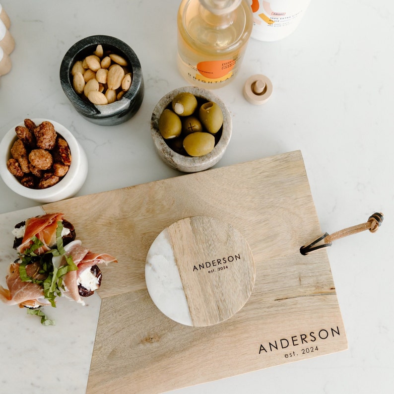Puede incluir: Una tabla de servir de madera con un posavasos de m&aacute;rmol y madera con el texto "Anderson est. 2024". La tabla tiene una asa de cuero y est&aacute; rodeada de cuencos de aceitunas, nueces y un aperitivo de queso y prosciutto.