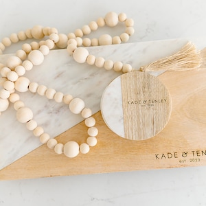 Puede incluir: Una tabla de cortar de madera con una incrustaci&oacute;n de m&aacute;rmol. La tabla tiene un posavasos redondo de madera y m&aacute;rmol con el texto "KADE & TENLEY est. 2023" grabado en &eacute;l. Una hebra de cuentas de madera est&aacute; encima de la tabla.