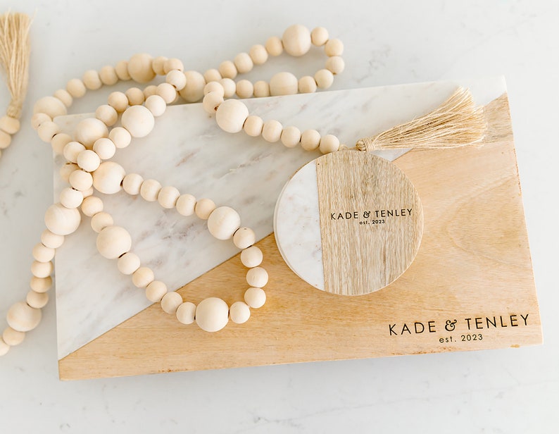 Puede incluir: Una tabla de cortar de madera con una incrustaci&oacute;n de m&aacute;rmol. La tabla tiene un posavasos redondo de madera y m&aacute;rmol con el texto "KADE & TENLEY est. 2023" grabado en &eacute;l. Una hebra de cuentas de madera est&aacute; encima de la tabla.