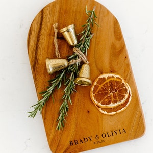 May include: A wooden cutting board with a rounded top, featuring the engraved text "BRADY & OLIVIA 6.22.23". Adorned with three small gold bells tied with twine, sprigs of rosemary, and dried orange slices. The board has a warm, natural wood grain.