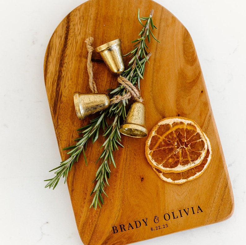 May include: A wooden cutting board with a rounded top, featuring the engraved text "BRADY & OLIVIA 6.22.23". Adorned with three small gold bells tied with twine, sprigs of rosemary, and dried orange slices. The board has a warm, natural wood grain.