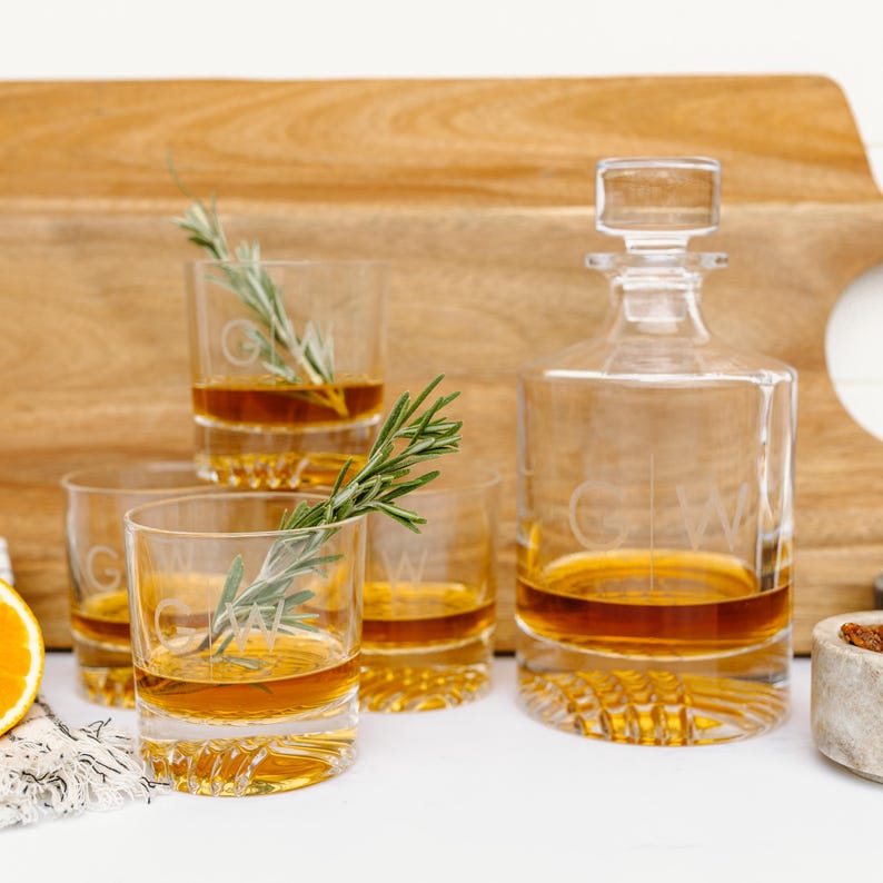 May include: Four clear glass whiskey glasses with a decorative etched design and a clear glass decanter with a stopper. The glasses and decanter are filled with a golden brown liquid. A sprig of rosemary is placed in one of the glasses.