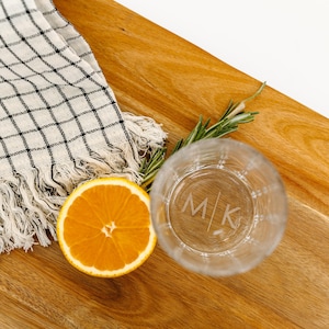May include: A clear glass with a textured surface and the initials "M|K" etched on the front. The glass is sitting on a wooden surface with a half orange and a sprig of rosemary.