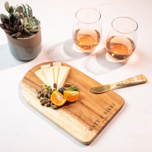 May include: A wooden serving board with cheese, pistachios, and a sliced orange. Two stemless wine glasses with a rose-colored liquid are in the background. A small succulent plant in a ceramic pot is also present. The board is engraved with "MATT & NIKKI est. 2019".