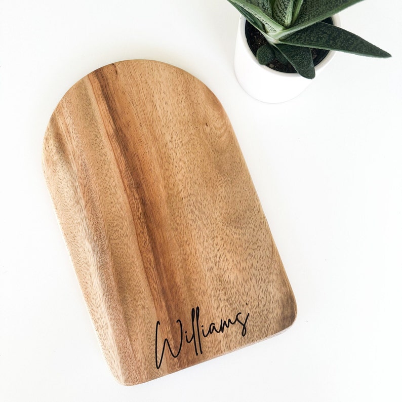 May include: A wooden arched cutting board with the name "Williams" in a cursive font. The board is light brown with a natural wood grain. A small potted succulent plant is in the background.