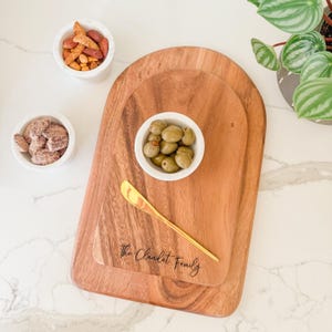 May include: Wooden serving boards with a gold cheese knife and small white bowls filled with snacks. The boards are engraved with "The Claudet Family". Olives, nuts, and other snacks are in the bowls. A potted plant is in the background.