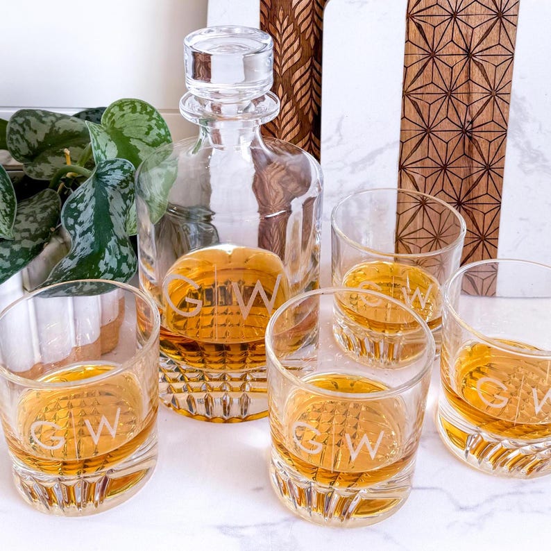 May include: A set of five clear glass decanter and glasses with etched initials "G.W." on each piece. The decanter has a round body and a stopper. The glasses are short and wide. The set is on a white marble surface.