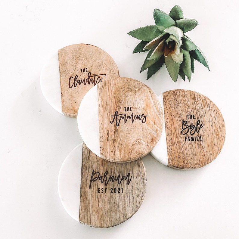 Puede incluir: Cuatro posavasos redondos de madera con incrustaciones de m&aacute;rmol blanco. Cada posavasos presenta un nombre de familia diferente en una fuente manuscrita. Los nombres son "The Claudats", "The Armons", "The Bogle Family" y "Parnum Est 2021".