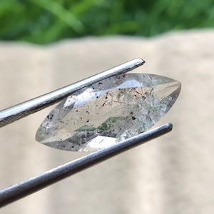 以下が含まれることがあります： 銀色のピンセットで挟まれた、透明なマーキス型の宝石。 宝石には赤、黒、緑の内包物があります。 背景はぼやけています。 宝石の長さは約2.5cmです。