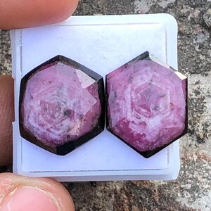 May include: Two pink and black gemstone cabochons with a hexagonal shape. The gemstones have a unique pattern of white lines and swirls.