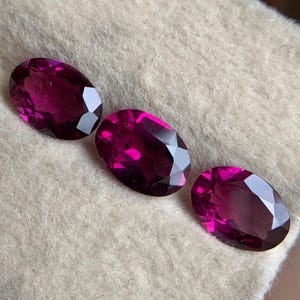 May include: Three oval-cut, faceted gemstones in varying shades of deep magenta and purplish-red. The gems are arranged on a light beige, textured surface, showcasing their polished facets and internal brilliance. The stones are likely garnets.