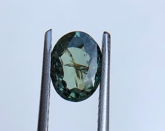 May include: An oval-shaped, teal gemstone is held by a pair of silver tweezers against a white background. The gem has internal inclusions and a multifaceted surface, reflecting light. The tweezers are positioned vertically, framing the gem.