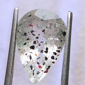 以下が含まれることがあります： 金属製のピンセットで保持された、洋梨型の宝石。 透明な石には、黒と赤のインクルージョンが散りばめられています。 宝石はカットされており、光を捉え、微妙な輝きを放ちます。 全体的な外観は半透明です。