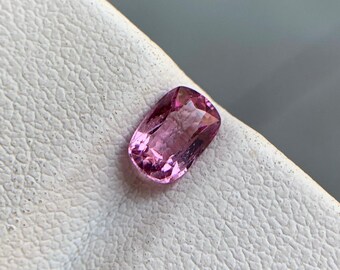 Op de afbeelding: Een gefacetteerde, ovaalvormige roze edelsteen. De edelsteen heeft een gepolijst oppervlak met meerdere facetten die licht reflecteren. De kleur is een levendig roze, met variaties in tint over de hele steen. De edelsteen ligt op een wit, getextureerd oppervlak.
