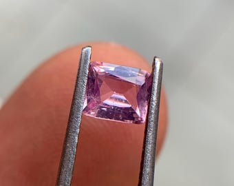 May include: A square-cut, light pink gemstone held by silver tweezers. The gemstone has a faceted surface that reflects light, showcasing its internal structure. The gem is a soft, pastel pink colour.