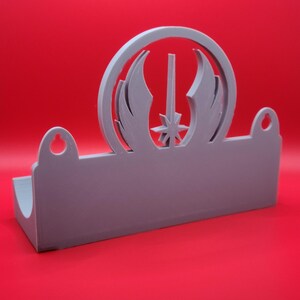 Puede incluir: Un soporte de pared de pl&aacute;stico gris con un recorte del s&iacute;mbolo de la Alianza Rebelde de Star Wars. El soporte tiene dos agujeros para colgar.