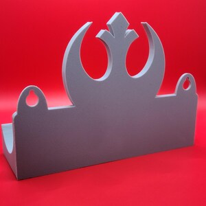 May include: A gray wall shelf with a Rebel Alliance symbol cut out. The shelf has two small holes on the sides.