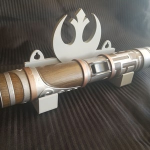 Puede incluir: Un soporte de pared blanco con un s&iacute;mbolo de la Alianza Rebelde y un sable de luz marr&oacute;n y plateado. El sable de luz tiene un dise&ntilde;o texturizado y un acento de cobre.