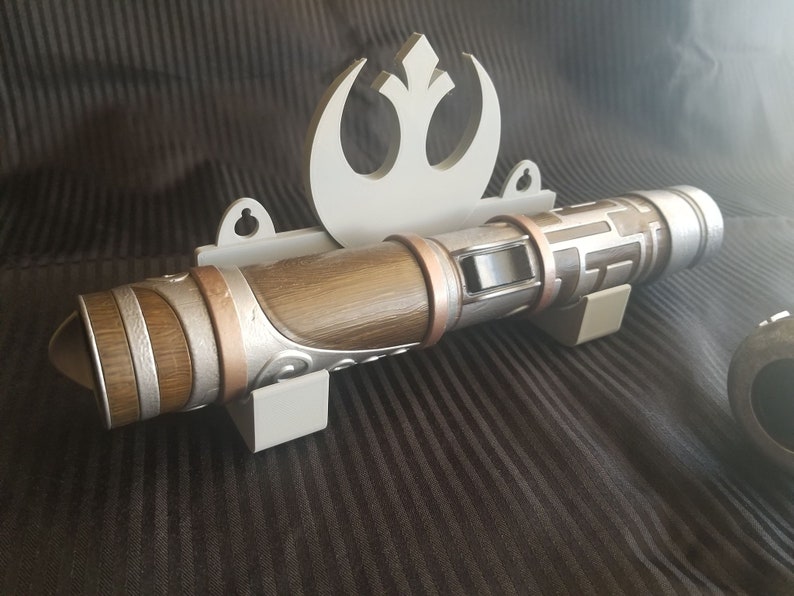 Puede incluir: Un soporte de pared blanco con un s&iacute;mbolo de la Alianza Rebelde y un sable de luz marr&oacute;n y plateado. El sable de luz tiene un dise&ntilde;o texturizado y un acento de cobre.