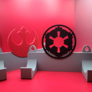 Puede incluir: Un estante de almacenamiento de pared con dos ganchos, que presenta un s&iacute;mbolo rojo de la Alianza Rebelde y un s&iacute;mbolo negro imperial. El estante est&aacute; hecho de pl&aacute;stico gris claro.