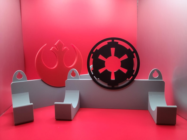 Puede incluir: Un estante de almacenamiento de pared con dos ganchos, que presenta un s&iacute;mbolo rojo de la Alianza Rebelde y un s&iacute;mbolo negro imperial. El estante est&aacute; hecho de pl&aacute;stico gris claro.