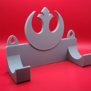 May include: A gray plastic wall mount with a Rebel Alliance symbol in the center. The mount has two hooks on the sides for hanging items.