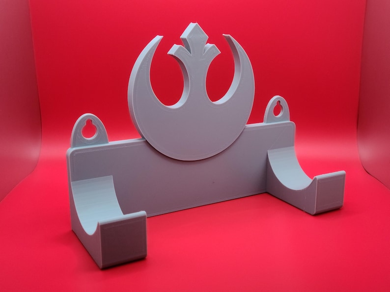 May include: A gray plastic wall mount with a Rebel Alliance symbol in the center. The mount has two hooks on the sides for hanging items.