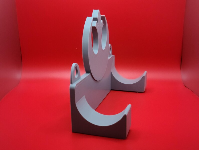 May include: A gray plastic wall mount with a Rebel Alliance symbol and two curved slots for holding items.