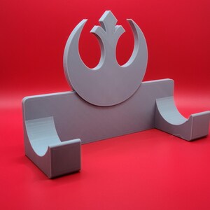 May include: A gray 3D printed wall mount with a Rebel Alliance symbol. The mount has two curved slots for holding items.