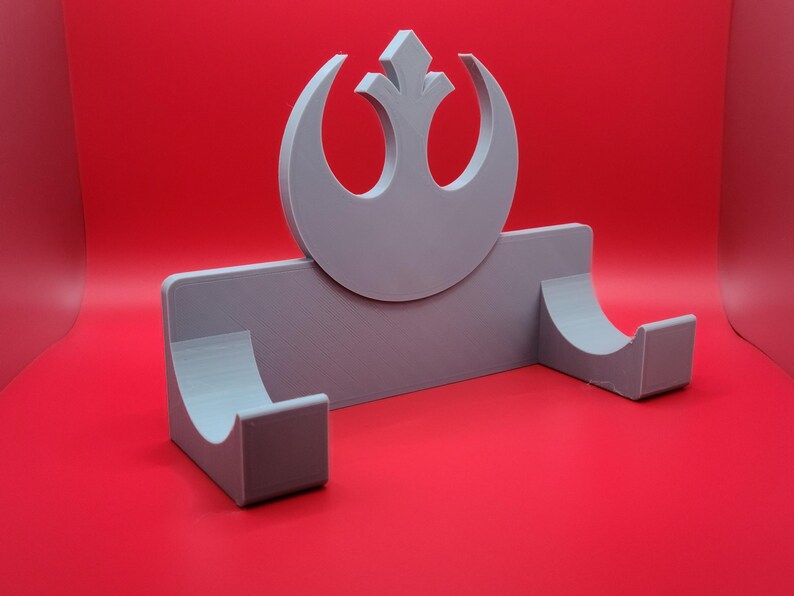 May include: A gray 3D printed wall mount with a Rebel Alliance symbol. The mount has two curved slots for holding items.