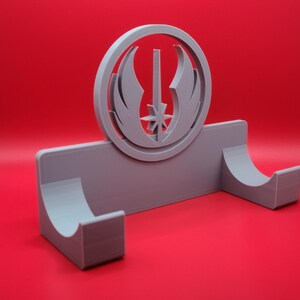 Puede incluir: Un soporte de pared gris impreso en 3D con un s&iacute;mbolo circular de la Orden Jedi en el centro. El soporte tiene dos brazos curvos para sujetar objetos.