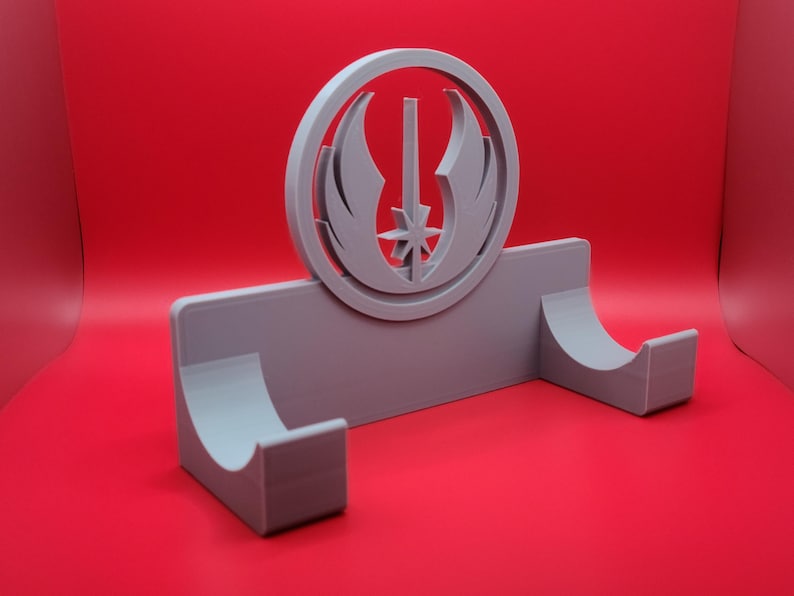 Puede incluir: Un soporte de pared gris impreso en 3D con un s&iacute;mbolo circular de la Orden Jedi en el centro. El soporte tiene dos brazos curvos para sujetar objetos.