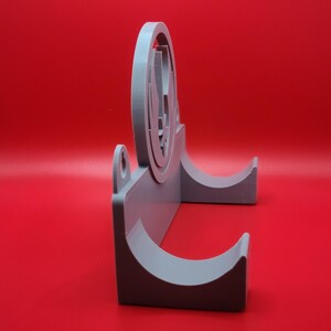 Puede incluir: Un soporte de pared de pl&aacute;stico gris con dos ganchos curvos. El soporte presenta un dise&ntilde;o circular en relieve con un s&iacute;mbolo estilizado.