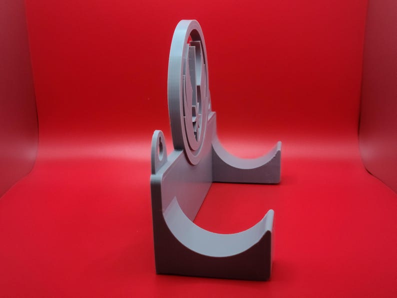 Puede incluir: Un soporte de pared de pl&aacute;stico gris con dos ganchos curvos. El soporte presenta un dise&ntilde;o circular en relieve con un s&iacute;mbolo estilizado.