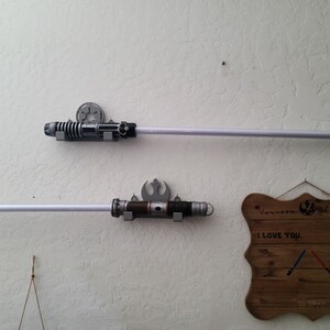 Puede incluir: Dos soportes de pared para sables de luz con sables de luz unidos. Los soportes est&aacute;n impresos en 3D y tienen un tema de Star Wars. Un letrero de madera con el texto "Te amo. Lo s&eacute;." est&aacute; colgado en la pared.