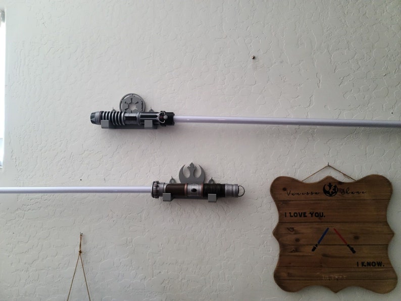 Puede incluir: Dos soportes de pared para sables de luz con sables de luz unidos. Los soportes est&aacute;n impresos en 3D y tienen un tema de Star Wars. Un letrero de madera con el texto "Te amo. Lo s&eacute;." est&aacute; colgado en la pared.