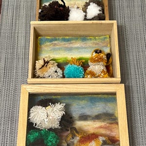May include: Three wooden shadow boxes with glass fronts, each containing a different scene of felted animal figures. The top box features three white, brown, and gray felted animals. The middle box features three felted animals, one brown, one blue, and one orange. The bottom box features two felted animals, one green and one orange, and a small sign that reads "FUTE".