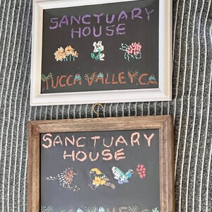 May include: Two framed signs with black backgrounds and white embroidery. The top sign says "Sanctuary House Yucca Valley, CA" and features embroidered flowers. The bottom sign says "Sanctuary House Yucca Valley, CA" and features embroidered birds.