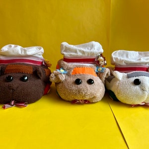 May include: Three handmade plush toys shaped like animals wearing white chef hats with red and white striped bands. The toys are made from brown, beige, and white fabric.