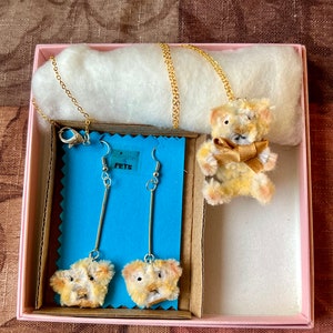 May include: A pink gift box with a white cotton lining contains a gold chain necklace with a fuzzy teddy bear charm, and a pair of dangle earrings with fuzzy teddy bear charms. The earrings are on blue cards with the word "PETS" printed on them.