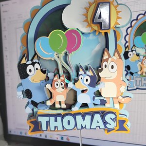 Op de afbeelding: Een blauwe, oranje en gele taarttopper met de personages uit de cartoon Bluey. De topper bevat een wolk met een zon en het nummer 4, drie ballonnen en de personages Bluey, Bingo en Bandit. De topper bevat ook een banner met de naam "THOMAS".