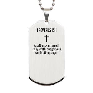 May include: A silver-coloured dog tag pendant with a ball chain necklace. The tag features the text "PROVERBS 15:1" above a cross symbol and a Bible verse: "A soft answer turneth away wrath: but grievous words stir up anger."