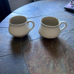 May include: Two off-white ceramic mugs with rounded bodies and small handles. Each mug has a thin, gold-colored band near the base. The mugs are sitting on a dark, textured surface. The mugs are likely for coffee or tea.