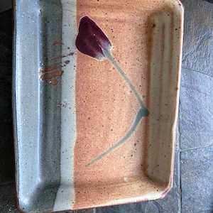 May include: A rectangular ceramic tray with a speckled finish, featuring a color-blocked design in shades of tan, cream, and muted green. A single dark maroon flower illustration with a green stem is painted in the center of the tray.