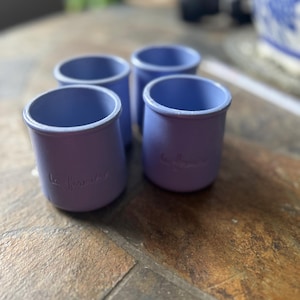 May include: Four small, cylindrical, lavender-colored ceramic cups with a slightly flared rim. The cups have the text "la fermière" in cursive on the side. The cups are arranged on a textured, brown surface.