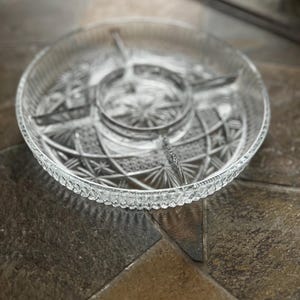May include: A clear, round glass serving dish with a starburst and geometric pattern. The dish is divided into sections, with a central circular compartment and radiating segments. The rim has a scalloped edge.