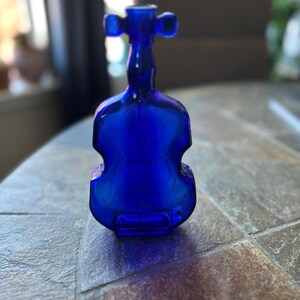 May include: A cobalt blue glass bottle in the shape of a violin. The bottle has a detailed design and a unique shape. The bottle is sitting on a stone surface.